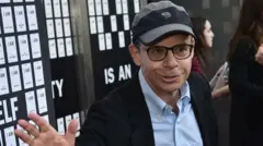 Rick Moranis usa óculos, boné preto, terno preto e camisa azul clara