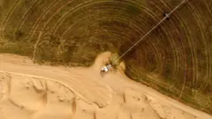 Вид с высоты на круглое зеленое сельскохозяйственное поле, граничащее с пустынным песком, с длинным трубопроводом, идущим от небольшого белого сооружения к зеленой зоне.