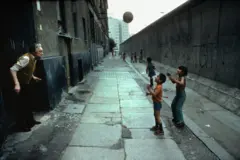 Crianças jogam futebol em uma rua com o Muro de Berlim passando pelo meio, impedindo o acesso ao outro lado, no Leste. Um homem idoso está parado ao lado de um prédio de tijolos que corre paralelamente ao muro.