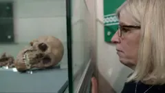 Uma mulher de óculos e cabelos loiros na altura dos ombros observa atentamente uma vitrine onde está exposto o esqueleto de um homem.