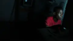 Um jovem aparece sentado diante de uma tela em um ambiente escuro, iluminado principalmente pela luz azulada do monitor que destaca seu rosto concentrado. Vestindo uma camiseta vermelha, ele olha fixamente para a tela enquanto utiliza um computador, cercado por um espaço pouco iluminado e com poucos detalhes visíveis ao fundo.