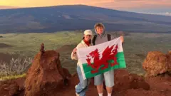 Hannah e Jack estão juntos, segurando a bandeira do País de Gales. Eles estão em frente a um campo verde e uma grande montanha. O céu está com uma coloração alaranjada e amarelada enquanto o sol começa a se pôr.