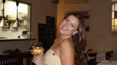 تقف ليبي ويلسون، البالغة من العمر 27 عامًا، مبتسمةً وهي تنظر إلى الكاميرا من جانبها. لديها شعر طويل بني فاتح ينسدل على ظهرها، وترتدي فستاناً أصفر فاتحاً منقطاً بنقاط بنية. إنها في مطعم، وتحمل كأساً من النبيذ.