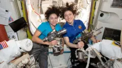 As astronautas Jessica Meir e Christina Koch flutuam lado a lado dentro da estação espacial, com os cabelos ao vento devido à ausência de gravidade. Jessica, à esquerda, com uma camiseta turquesa, e Christina, à direita, com uma camisa polo azul, seguram cada uma uma grande ferramenta elétrica prateada usada para manutenção dos trajes espaciais. Ao redor delas, peças brancas dos trajes espaciais, cabos e painéis criam uma atmosfera de oficina movimentada dentro do interior curvo e claro do módulo da câmara de descompressão