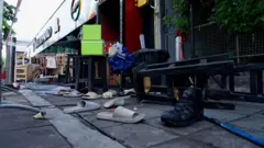 Pares de chinelos levemente chamuscados espalhados na rua, do lado de fora do bar, com banquetas altas ao fundo