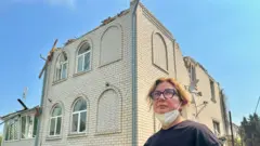 れんが造りの一軒家の屋根部分が吹き飛び、外壁にも損傷が見られる。家の外には、マスクをあごにかけた女性が立っている