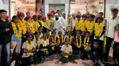Uma equipe feminina de futebol aparece vestida com roupas esportivas e usando pompons amarelos. Dois homens que estão entre elas seguram troféus