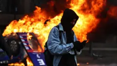 Una persona con el rostro cubierto mira su teléfono mientras arde un auto en las protestas en Buenos Aires. 