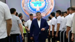 Oq ko‘ylak va to‘q ko‘k kostyum-shim kiygan Qamchibek Tashiev oq ko‘ylakdagi erkak amaldorlar orasida yurib ketmoqda.