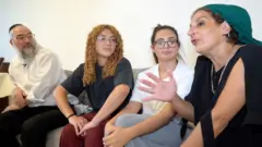 Quatro pessoas estão sentadas em um sofá: um homem de camisa branca, quipá e óculos; uma jovem de cabelo castanho-claro encaracolado, longo, usando óculos, camiseta preta e calça cor terracota; uma jovem de cabelo escuro liso preso para trás, usando óculos, camiseta branca e calça cinza; e uma mulher mais velha com camiseta preta e lenço verde na cabeça.