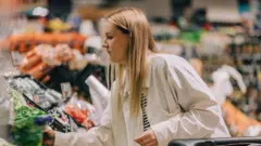 Kobieta o blond włosach wybiera warzywa w supermarkecie, ma na sobie białą koszulę