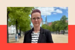 Una mujer blanca con el pelo morado recogido hacia atrás y gafas. Lleva una americana oscura y una camiseta de rayas blancas y negras, y mira a la cámara