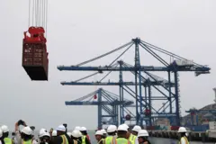 Grúas transportan un contenedor en el gran puerto de Chancay. Debajo, en el suelo, un grupo de personas con cascos y chalecos reflectantes.