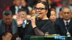 Pastora, segurando um microfone, sorri durante pregação em um púlpito, com pessoas desfocadas atrás. Ela veste preto, usa óculos e está com os cabelos presos.