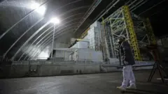 У великому закритому приміщенні з аркоподібною металевою стелею стоїть масивна промислова конструкція з бетону й металу, освітлена яскравими лампами. На передньому плані людина в захисному одязі, касці та масці повільно йде бетонною підлогою, поруч видно інженерне обладнання.