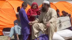 A bearded man wearing a beige shirt and trousers and a white hat sits on some sacks. He is holding the hand of a child who is wearing a purple shirt and trousers. A girl stands between them wearing a red headscarf. Another child in purple lies on the ground at the man's feet. There are orange tents and a chest behind them.