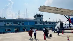 কক্সবাজার বিমানবন্দরের দুই তলা ভবন। সামনে একটি বিমানে উঠছেন কিছু যাত্রী যাদের মধ্যে একজন পুরুষের কোলে একটি শিশু রয়েছে এবং পুরুষটি এক নারীর হাত ধরে আছেন। বিমানের সামনের দিকে একটি দরজা খোলা এবং সেখানে ট্রলিতে করে মালপত্র তোলা হচ্ছে। 