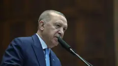 Lacivert çizgili takım elbise giymiş bir adam kürsüde konuşuyor