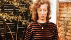 Foto antiga de Christine Harverson, com cabelo cacheado, está ao ar livre em frente a uma cerca de madeira e algumas plantas. Ela está usando uma camisa de manga longa de cor escura com listras horizontais e um colar com um pequeno pingente. Ao fundo, há vegetação e parte de uma parede de tijolos