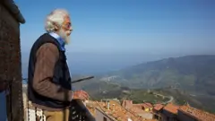 Homem idoso olha para a cidade e as montanhas do terraço de uma casa