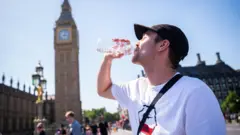 ロンドン・ウェストミンスター橋で、白いTシャツに黒いキャップ姿の男性が、ペットボトルの水を飲んでいる。背景に、ビッグベンや議事堂が並ぶ。