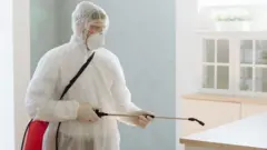 Foto arsip seorang kontraktor profesional mengenakan pakaian pelindung dan masker sedang menyemprotkan disinfektan untuk hama atau virus di sebuah rumah.