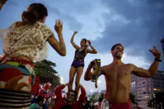 Pessoas dançam com os braços para a cima na praia. Um homem está sem camisa. Outro carrega uma mulher nos ombros.