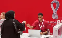 Un hombre sonriente con una camiseta roja ayuda a una mujer vestida de negro. Ella sostiene algo que parece una tarjeta de identificación con un cordón. La pared que hay detrás del hombre es de color rojo brillante.
