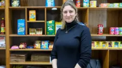 Narion Coelho, uma mulher loira de meia idade, vestindo uma blusa preta de gola rolê e sorrindo discretamente para a câmera, em frente a uma estante com produtos brasileiros, como erva-mate, café e macarrão instantâneo