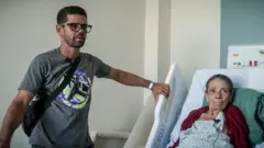 Una anciana en una acostada en una cama hospitalaria junto a su hijo.