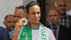 Imane Khelif sostiene una medalla. Hay varios hombres detrás de ella.