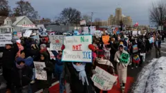 曇り空の下、防寒対策をした人たちが移民税関捜査局（ICE）に抗議するプラカードなどを手に行進している。沿道には雪が積みあがっている