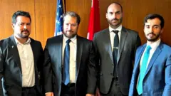 Quatro homens lado a lado em sala, posando para foto com feições contidas