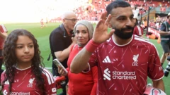 Muhammed Salah, yanında kızı ve eşiyle sahada yürüyor ve Liverpool taraftarına veda ediyor. Elini kaldırmış durumda