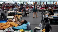 O femeie cu păr lung, închis la culoare, stă întinsă și se uită la ecranul telefonului, sâmbătă, la Centrul Expozițional Bordeaux-Lac, care a fost transformat într-un adăpost pentru persoanele evacuate. În fundal, se văd paturi și alte persoane.

