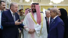 Cumhurbaşkanı Erdoğan solda, Muhammed bin Salman ortada, Şahbaz Şerif sağda. Üç lider yürürken gülüşerek sohbet ediyor. Etraflarında danışmanları ve askeri üniformalı kişiler var. Erdoğan ve Şerif takım elbise giyiyor, bin Salman'ın üzerinde geleneksel Suudi Arabistan kıyafeti var. Arka planda sarı renkli duvarlar görünüyor.