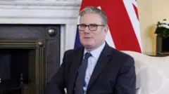 大理石と鉄細工でふちどられた暖炉の前、白い布張りの肘掛椅子に座ってカメラを見るスーツ姿のスターマー氏