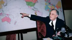 Roosevelt sentado em uma cadeira apontando para um mapa.