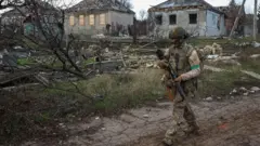 ukrajinski vojnik korača sa puškom u ruci, u pozadini su oštećene kuće