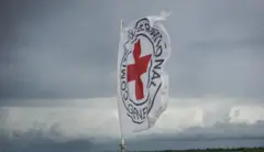 Ibendera ry'umweru rya CICR ririmo umusaraba utukura hamwe n'amagambo yandikishijwe ibara ry'umukara. Inyuma kure haraboneka ibicu by'ubururu n'umweru.