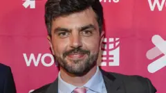Will Adolphy, um homem com barba e cabelo curto, vestido com terno, camisa azul e gravata rosa. Ele sorri olhando diretamente para a câmera.