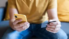 Una persona con una camiseta amarilla sostiene un celular del mismo color en una mano y una tarjeta de pago en la otra. 