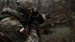 Żołnierz w kamuflażu z polską flagą na ramieniu i z karabinem szturmowym 