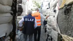 OLAF ve ARPAT yazılı yetkililer tekstik atıklarının arasından geçiyor, arkaları dönük. 