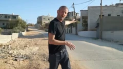 Periodista parado frente a la cámara enseñando casas en una calle de Cisjordania
