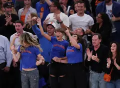 Taylor Swift aparece na primeira fila da quadra, ao lado de outras pessoas, torcendo pelo New York Knicks. Ela veste uma camiseta azul com letras laranja, nas cores do time