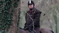 Wearing riding clothes, including a helmet, Andrew Mountbatten-Windsor is shown between the branches of a tree, riding a horse and looking beyond the camera. 