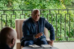 Joseph Kabila, ex-président de la RDC en costume bleu avec une chemise carrelée bleue et blanche, lève la main droite en s'adressant à des gens. 