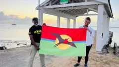 Os ativistas John Mussington e Jackie Frank seguram a bandeira de Barbuda à beira-mar