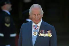 الملك تشارلز الثالث مرتدياً بدلة رسمية لونها بين الكحلي والأزرق مقلمة بلون أبيض وربطة عنق زرقاء منقطة وقميص أبيض. ومجموعة من الأوسمة على جاكيت بدلته وقلادة في رقبته.